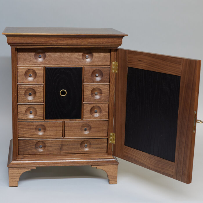 Ett handgjort kryddskåp av amerikansk 1700-talsmodell med en öppen dörr, flera små lådor och ett centralt skåp, tillverkat av trä med en mörk insats mitt i.