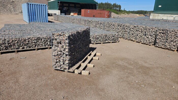Gabionburar fyllda med stenar står uppradade på en grusplan vid en byggarbetsplats, med containrar och en byggnad i bakgrunden.