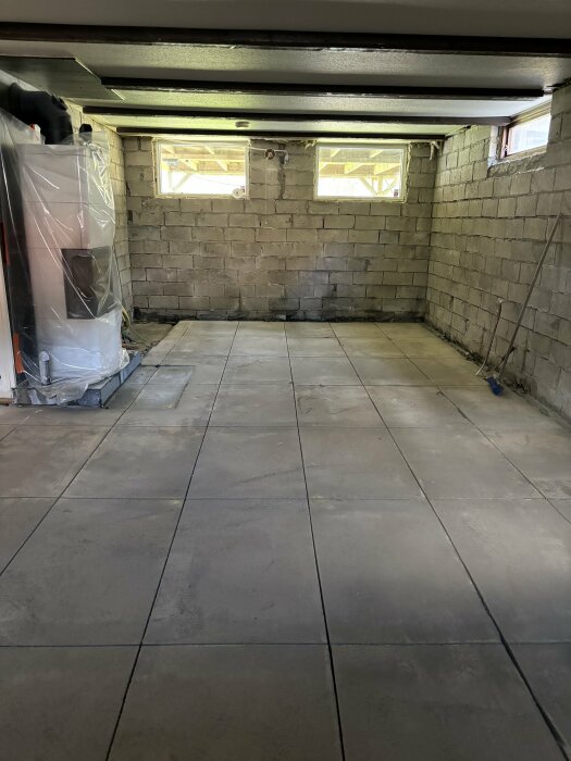 Källare med betonggolv innan renovering, två små fönster, tegelstensväggar och en täckt vit pannpanna till vänster. Golvet är täckt med plattor.