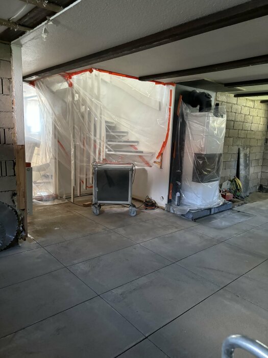 Gamla betonggolvplattor i en renoveringsprocess, täckta trappa med plastskynke i bakgrunden. Golvet är utjämnat med makadam.