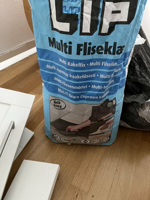 En påse med blå etikett som innehåller kakelfix med texten "Multi Fliseklæb" och "Grå Grey", står på ett trägolv med vita kakelplattor bredvid.