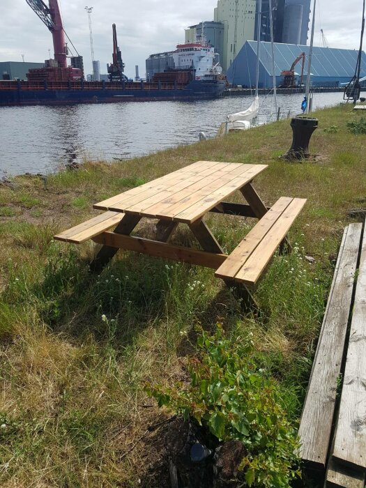 Träbord, byggt med återvunnet material, står vid en gräsmatta nära en hamn med fartyg i bakgrunden.