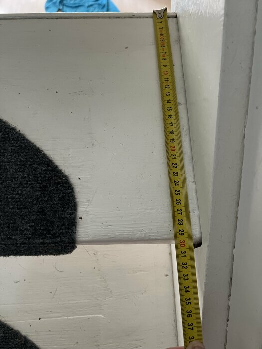 Närbild på en trapprenovering, ett trappsteg mäts med ett måttband som visar 300 mm bredd. Trappsteget är målat vitt och har grå mattor.
