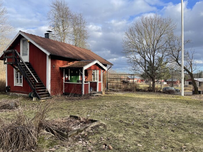 Rött torp med svart trappa och vitmålade fönsterkarmar, omgivet av gråa träd och vissna buskar. Glest molntäckt bakgrund och bil i fjärran.