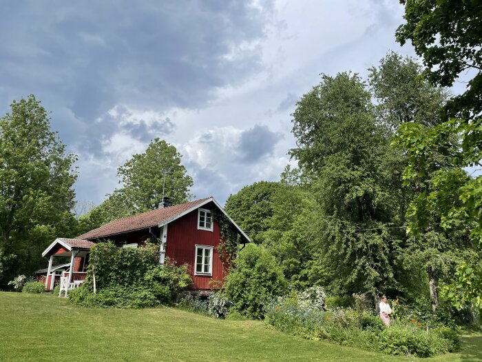 Rött torp med vita knutar, omlivet av frodig grönska och blommor, med mörka moln på himlen.