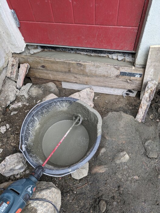 En nyligen gjuten betongyta under en röd källardörr, med en svart plastbehållare med flytande betong och en borrmaskin till vänster.