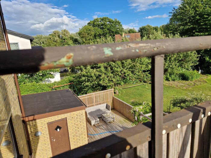 Balkongräcke av metall med rost och avskalad färg sett från en balkong med utsikt över en trädgård och en träterrass med utemöbler.