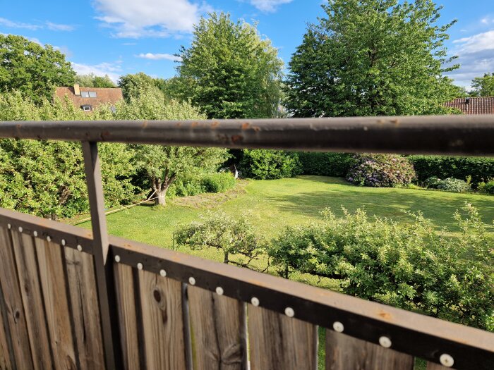 Rostigt metallräcke mot träpanel på balkong med utsikt över en frodig trädgård med gräsmatta, träd och buskar under en klar blå himmel.