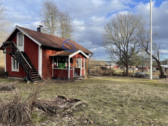 Rött torp med trappa på utsidan, mörkt tak och en genomskinlig dörr på övervåningen. Ett blått märke indikerar en läcka i taket. Träd och gårdsbyggnader syns i bakgrunden.