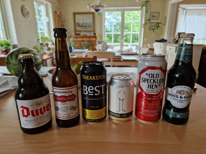 Sex olika öl på ett bord med en köksmiljö i bakgrunden; märken inkluderar Duvel, Göteborgs Nya Starkpilsner, Theakston Best, Old Speckled Hen och Innis & Gunn.