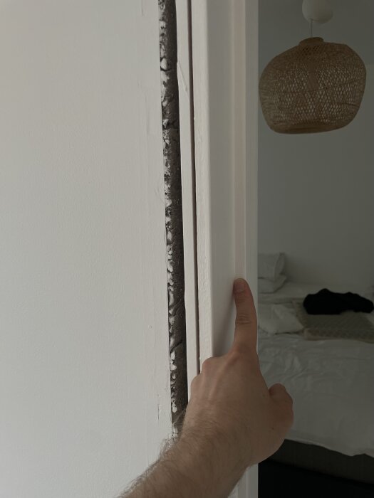En hand som trycker på en innerdörr för att visa dörrkarmen inifrån ett rum med en säng och en taklampa i bakgrunden.