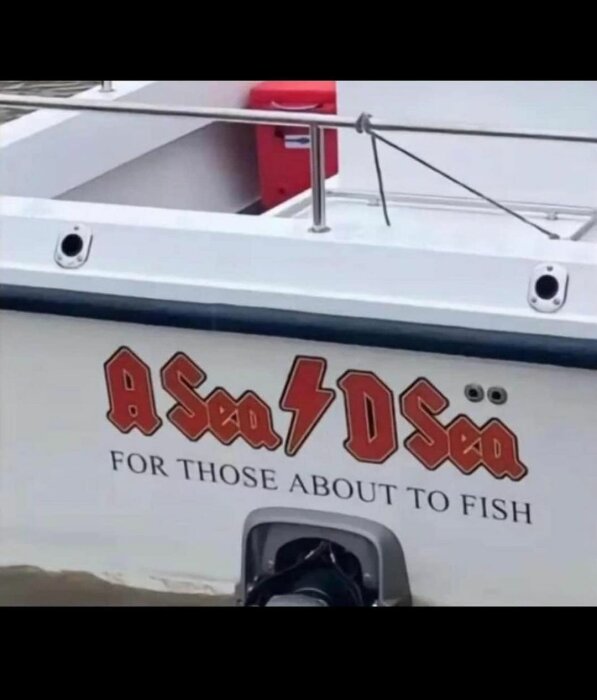 En båt med texten "A Sea D Sea" och "For those about to fish" på sidan, samt metallräcke och en röd förvaringslåda i bakgrunden.