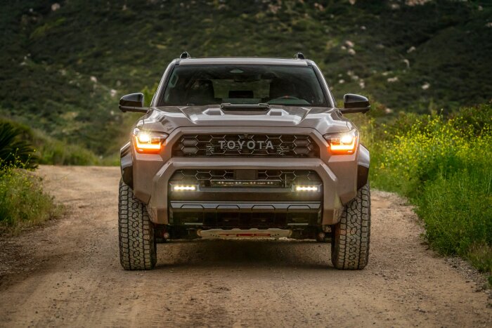 En ny Toyota SUV kör på en grusväg, fotograferad framifrån med tänd belysning och bergig natur i bakgrunden.