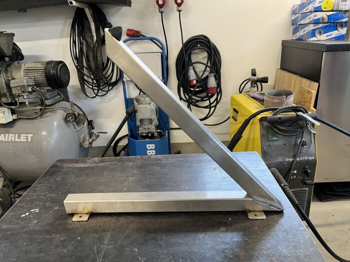 Självtillverkad dörrstopp i metall står på ett arbetsbord i en verkstad med olika verktyg och utrustning i bakgrunden.