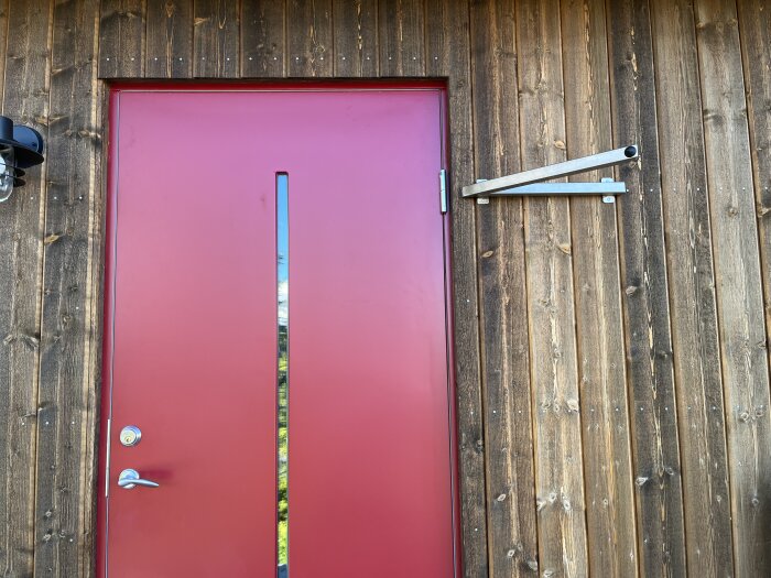 Röd ytterdörr i träfasad med nyinstallerat dörrstopp av metall monterat ovanför dörren. Dörrstoppet förhindrar att dörren blåser upp och orsakar skador.
