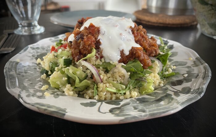 En tallrik med chili con carne och vitlökssås ovanpå en sallad med bulgur, grönsaker och lök.