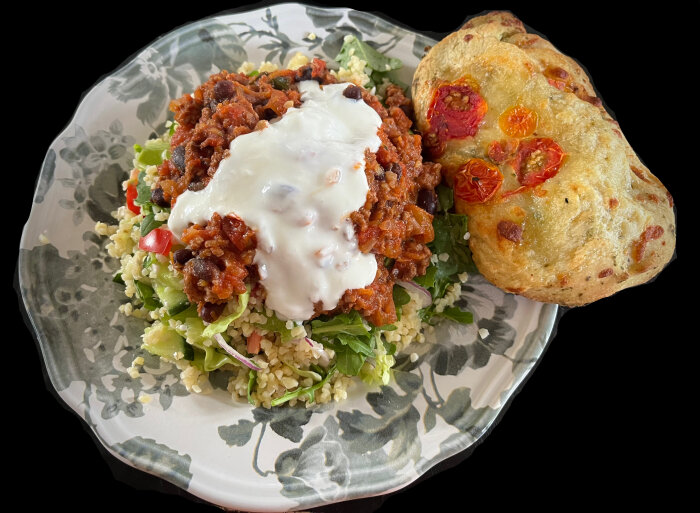 Tallrik med chili con carne, bulgursallad, vitlökssås och focaccia dekorerad med tomater.