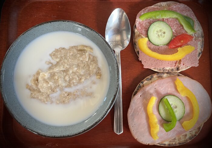 Skål med havregrynsgröt och mjölk, bredvid en sked och två rågmackor med köttpålägg, paprikabitar, gurkskivor, och tomatbit på en träbricka.