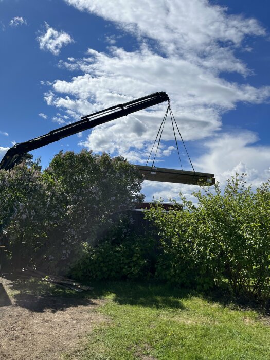 En kran lyfter en stor planka över en grönskande trädgård under en klarblå himmel med moln.