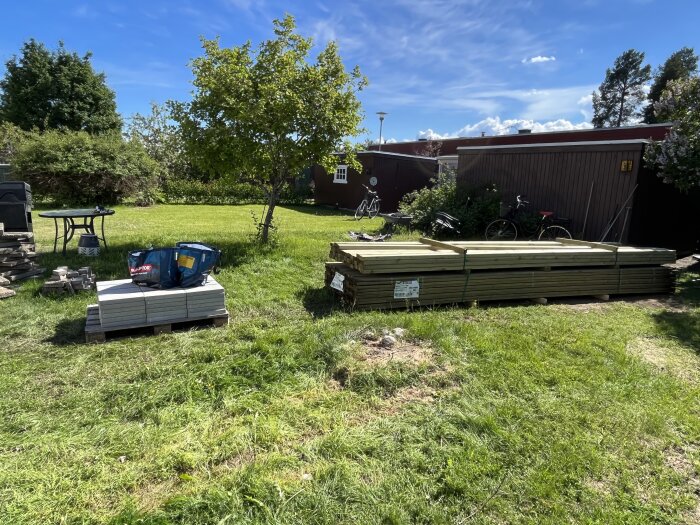 Gräsmatta med staplad byggmaterial som träplankor och stenplattor samt en blå verktygslåda. I bakgrunden syns ett träd, en trädgårdsbord och en cykel vid en byggnad.