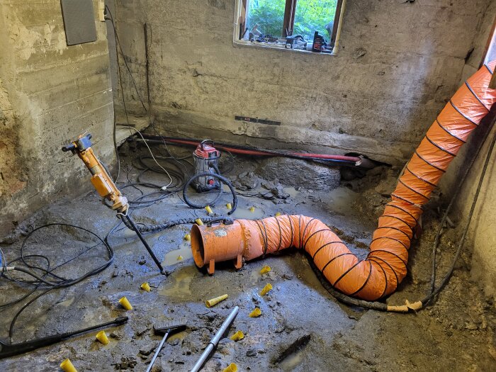 Borrutrustning och ventilationsslang i en betongvägg i ett rum med borrhål och spridda plastkoner. Arbe förberett för snigeldynamit.