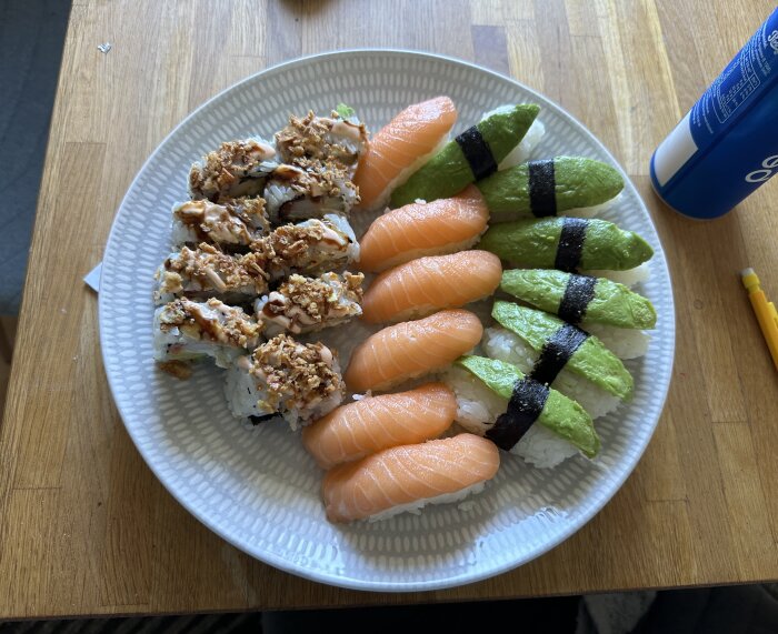 En tallrik med sushi, inklusive rullar toppade med krispig lök, lax nigiri och avokado nigiri, placerad på ett träbord med en dryck nära.