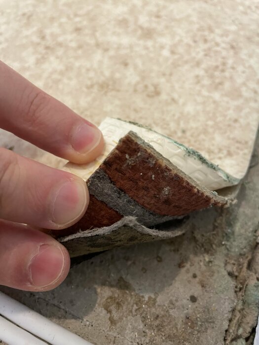 En hand håller upp en bit av en brun plastmatta med flera lager, som ser ut att vara gammal och sliten, mot en bakgrund av grusig betong.