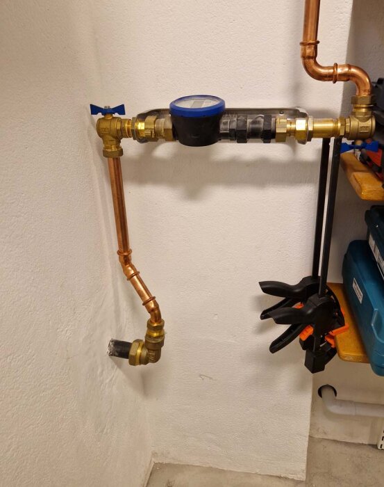 Kopparrör och vattenmätarkonsol installerade på en vägg i tvättstuga, med anslutningar och genomföring genom väggen, samt några monterade klämmor på hyllan intill.