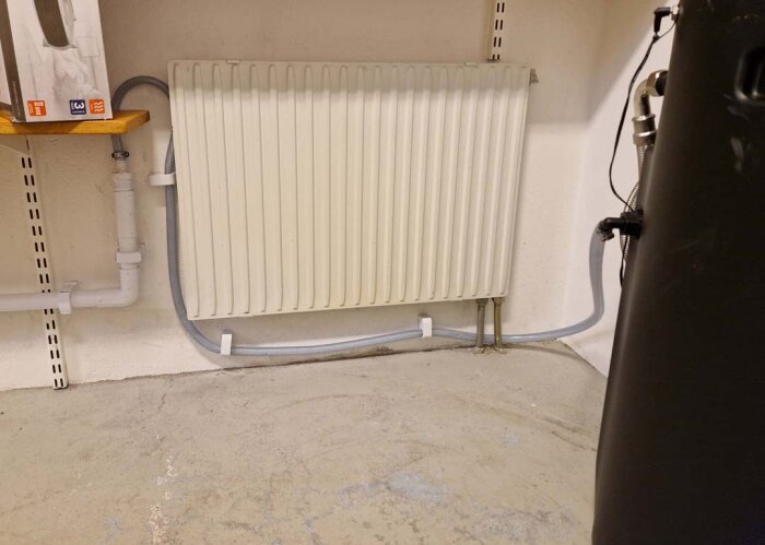 En vit radiator installerad på en vägg med tillhörande rördragningar och en hylla ovanför. Golvet är av betong och avlutningssystemet synligt.