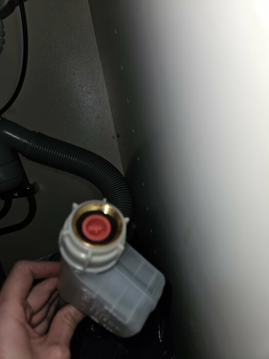 Hand holding a dishwasher connection hose with G3/8 socket, highlighted by a red inner part. Surrounding area includes pipes and part of a cabinet wall.