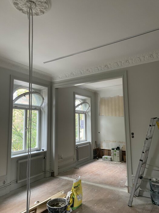 Bilden visar ett pågående renoveringsprojekt i ett rum med höga fönster och ljusgrå väggar. Golvet är oklart och byggmateriel, inklusive verktyg och färgburkar, syns på golvet och en stege står uppställd.