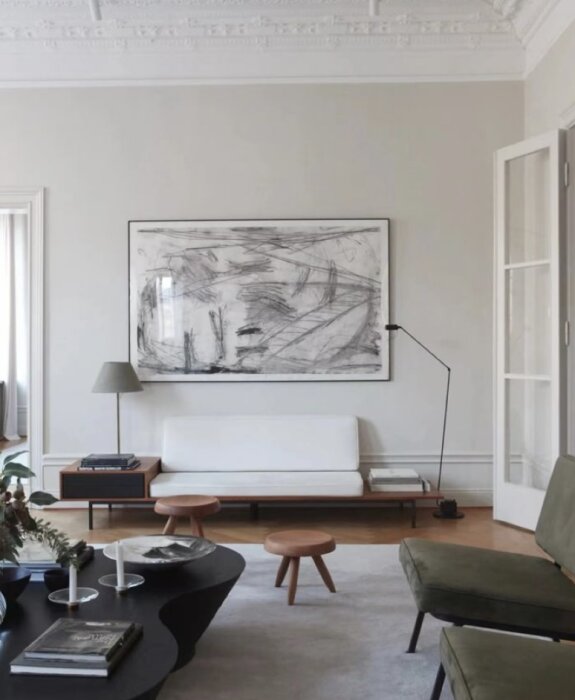 Modernt vardagsrum med ljusgrå väggar, vit soffa, svart-vitt abstrakt konstverk, mörkgrönt fåtölj och trätaburetter med golvlampa.