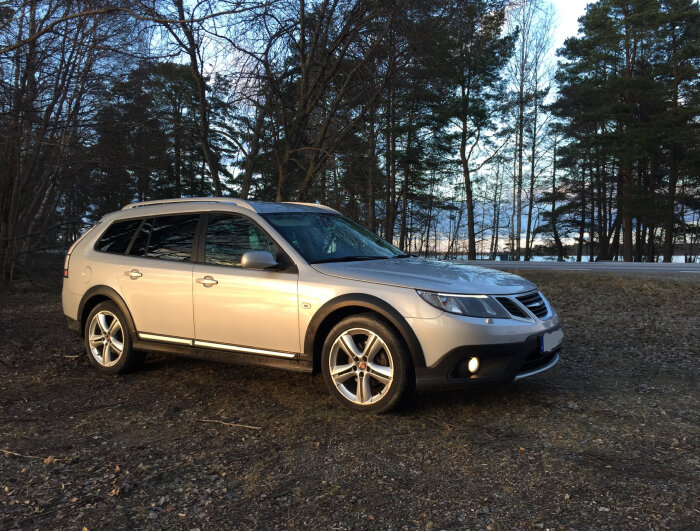 SIlverfärgad kombibil parkerad på en grusväg i skogsmiljö med träd i bakgrunden och disig himmel.