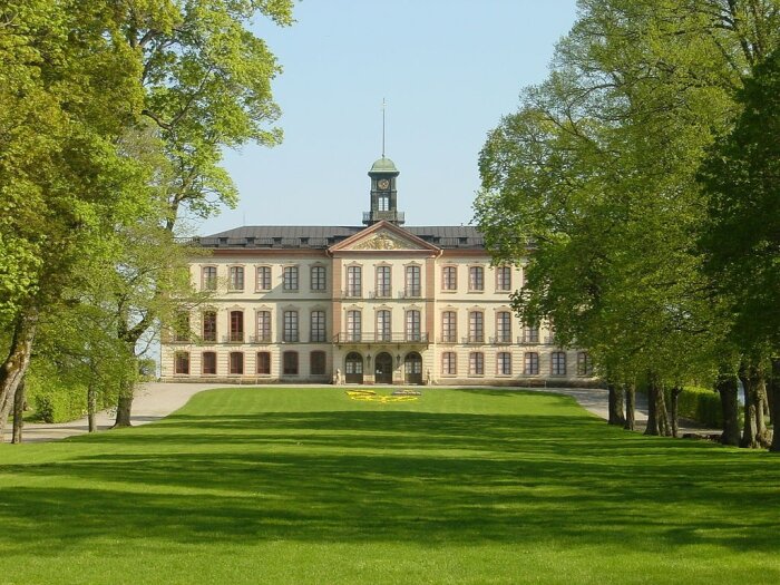 Tullgarns slott, en stor byggnad med flera fönster och en liten torn, omgiven av träd och grön gräsmatta.