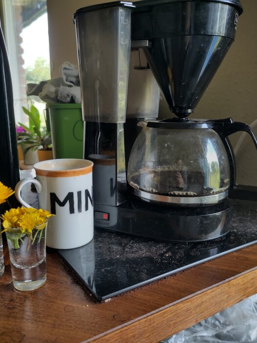 En kaffebryggare med Melitta-logotyp står på en bänk med en halvfull kaffekanna. Bredvid står en kopp märkt "MIN" och en liten vas med gula blommor.