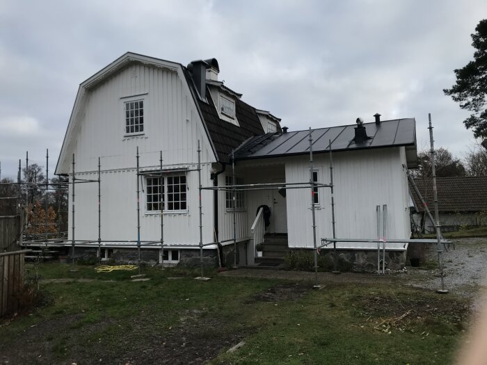 Vit tvåvåningsbyggnad med byggnadsställningar runtom, grått tak och fönster, gräsmatta och grusgång framför, träd och buskar i bakgrunden.