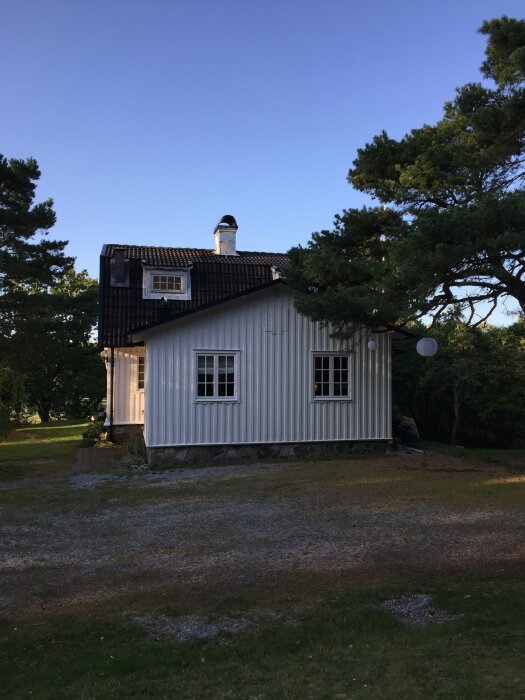 Vitt tvåvåningshus med brunt tak och två fönster på första våningen, omgiven av grönska och träd, med klar blå himmel i bakgrunden.