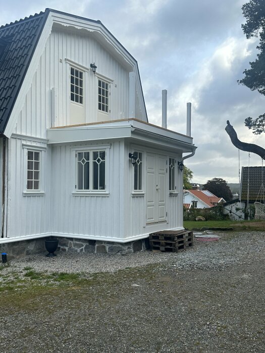 Byggnad med vit fasad och svart tak, två utskjutande rör från en balkong, tre fönster på bottenvåningen och två på övre våningen, vid en grusad gårdsplan.