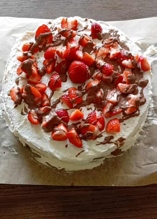 En tårta täckt med vispgrädde, toppad med färska jordgubbar och ringlad choklad, placerad på ett bakplåtspapper på en träyta.