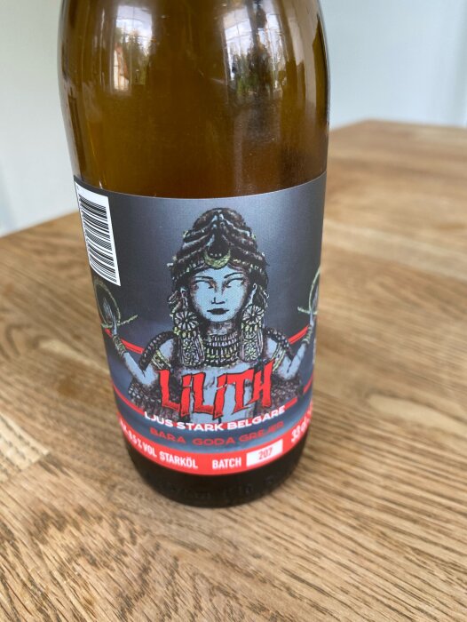 En flaska ölet "Lilith" från Snausarve gårdsbryggeri, ljus belgisk stil med etikett som avbildar en figur och texten "Bara goda grejer". Placerad på träbord.