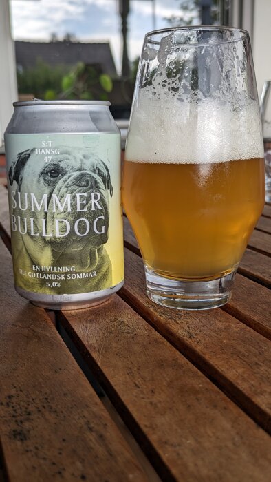 En burk med öl med etiketten "Summer Bulldog" bredvid ett glas fyllt med skummande öl, stående på ett träbord utomhus.