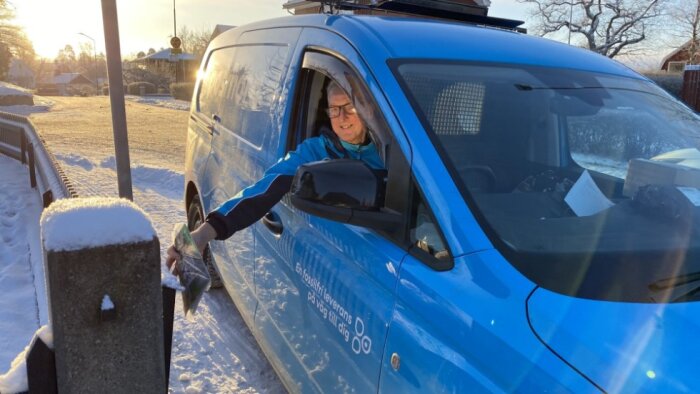 En postanställd i en blå skåpbil delar ut post i en brevlåda från höger hand genom fordonets fönster på en snötäckt uppfart.