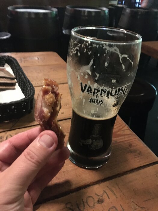 Hand som håller i ett grisöra bredvid ett glas med mörkt öl som är märkt "Varniuku" men som egentligen innehåller Pekla (4,2%), bord och fat i bakgrunden.