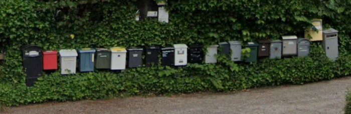 En rad med brevlådor i olika färger och storlekar monterade på en grön häck, ingen av brevlådorna är i anslutning till ett hus.