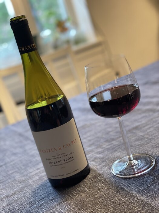 En flaska rött vin av märket Frantzén & Cauble och ett glas rödvin stående på ett grått bord.