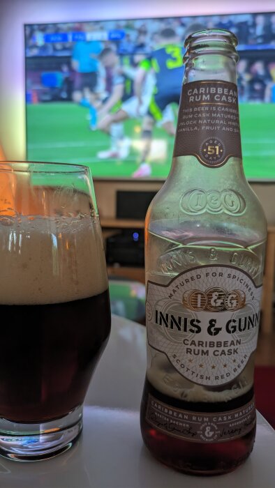 En flaska Innis & Gunn Caribbean Rum Cask öl och ett fyllt ölglas i förgrunden, en fotbollsmatch visas på en TV i bakgrunden.