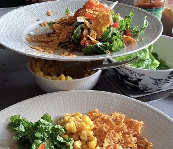 Två tallrikar med tacorätter redo att ätas. Den övre tallriken innehåller lammfärs, sallad, tomater och Doritos. Den nedre tallriken har sallad, majs och Doritos.