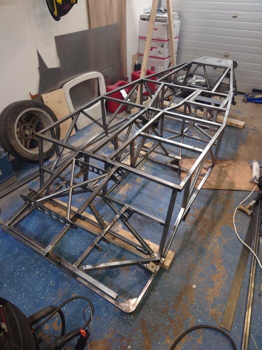 En metallram som del av ett byggprojekt ligger uppochned i ett garage, omgiven av verktyg och material som däck, brädor och metallplattor.