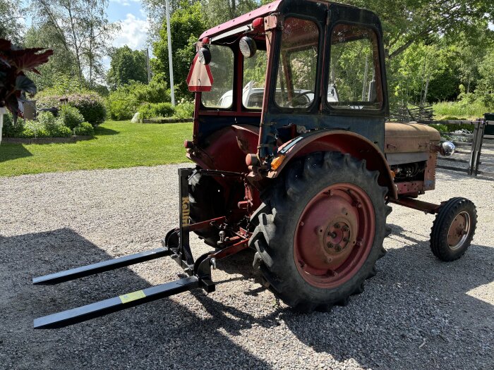 Röd traktor på en grusplan, med skylt bakom föraren och svarta gafflar monterade bak för att lyfta och flytta föremål. Grönskande trädgård i bakgrunden.