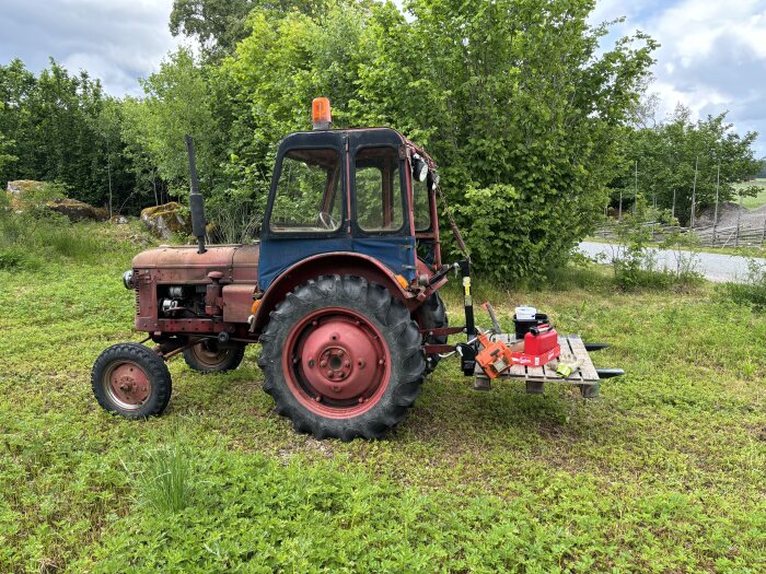 En gammal traktor med bakmonterade gafflar lyfter en pall med olika prylar inklusive en motorsåg och en verktygslåda på en gräsmatta med träd i bakgrunden.